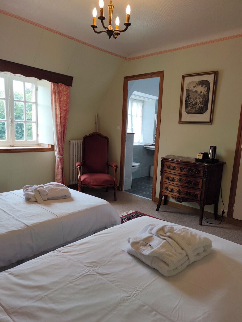 Camera d'albergo con due letti, una poltrona rossa, una cassettiera e un lampadario.