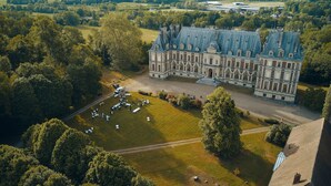 An aerial view of a large, historic building with a symmetrical facade, a central courtyard, and a surrounding lush green landscape.