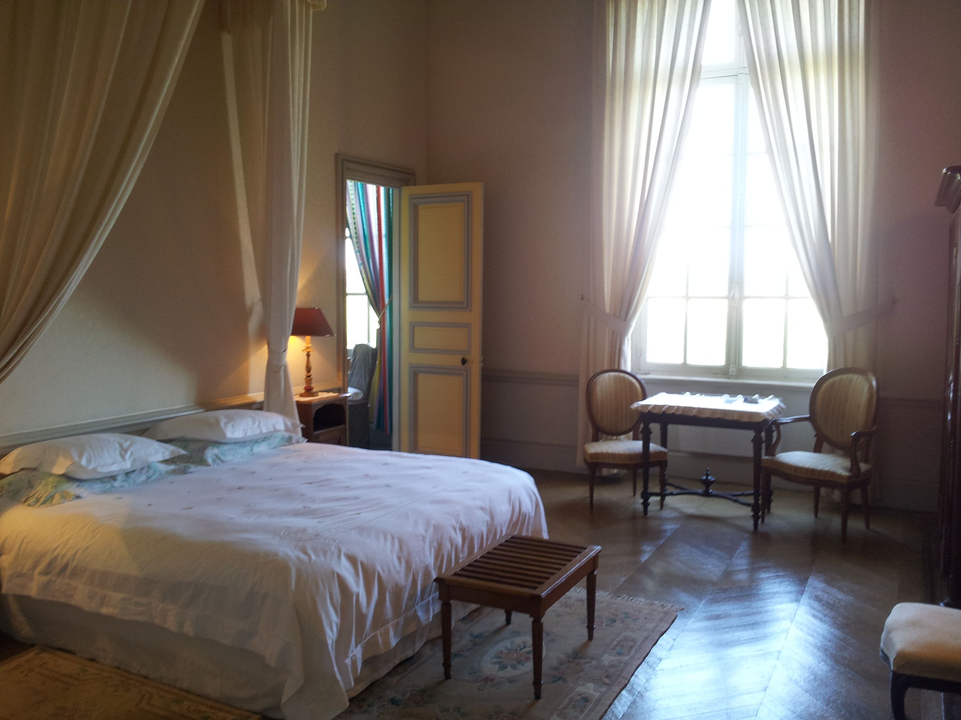 A bedroom with a large bed, a wooden table, two chairs, a desk, and a window with curtains.
