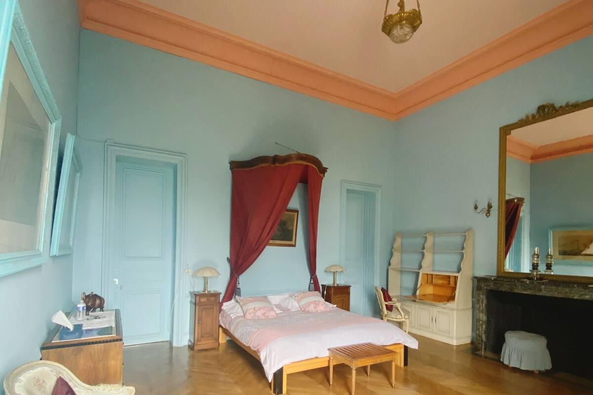 Quarto com piso de madeira, cama com dossel, uma cadeira, cômoda e espelho.