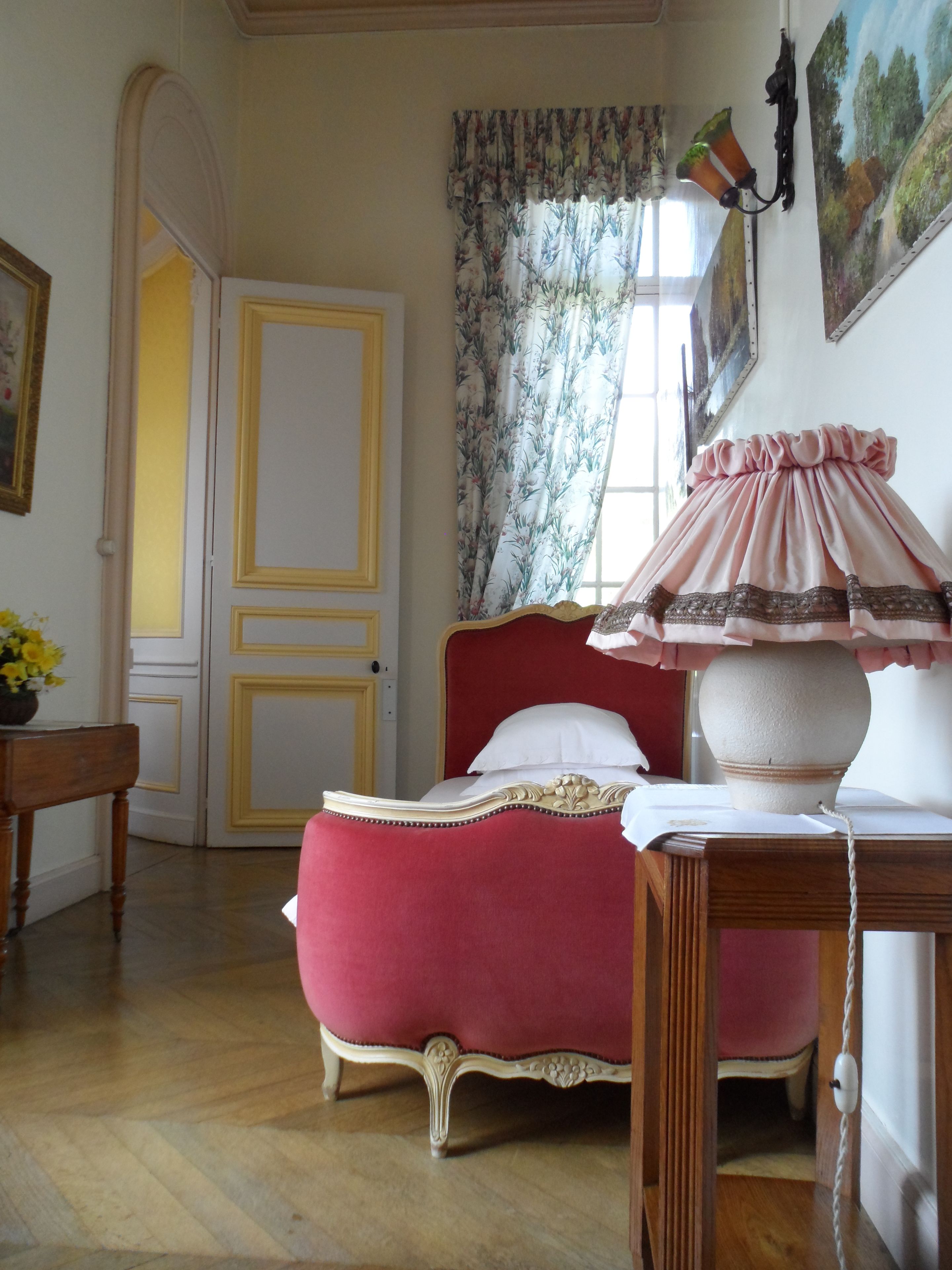Quarto com cama, mesa e abajur. Há uma porta e uma janela com cortinas.