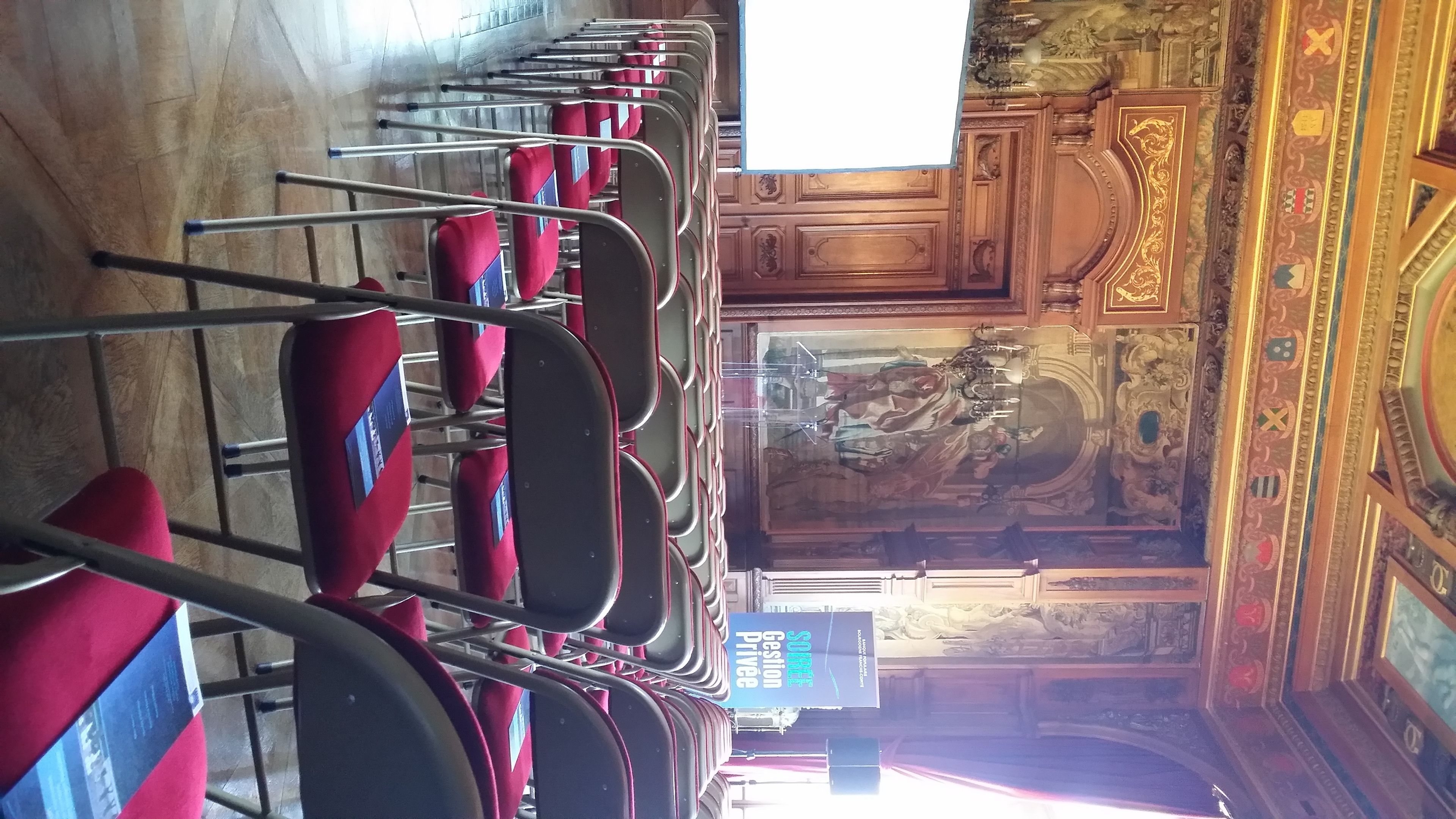 Des rangées de chaises rouges à armature métallique, une enseigne indiquant « SODERE Gestion Privée », et un décor mural en bois ouvragé avec une fresque en arrière-plan.