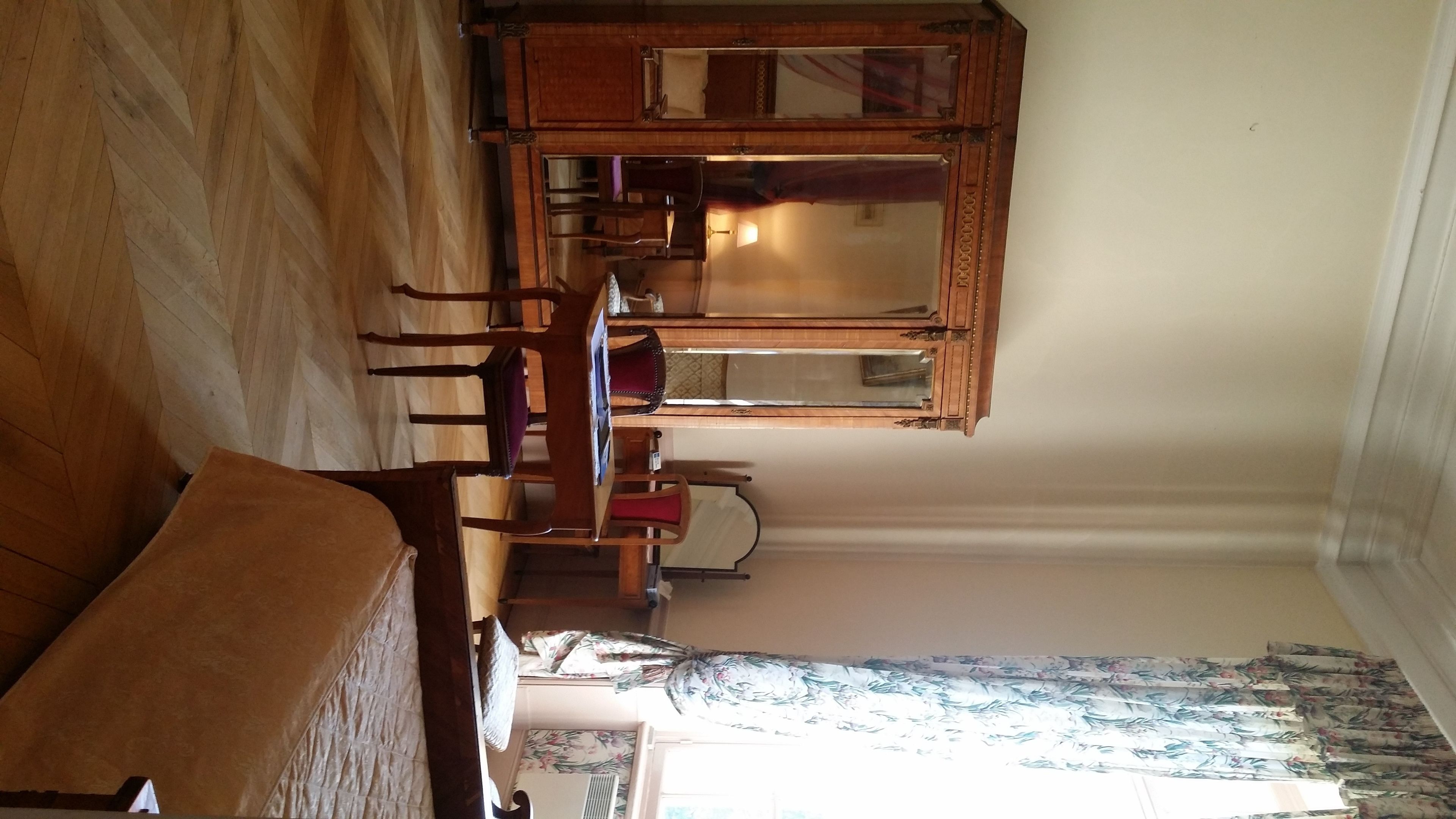 A room with a wooden floor, a wooden dining table with chairs, a mirror, and a window with floral curtains.