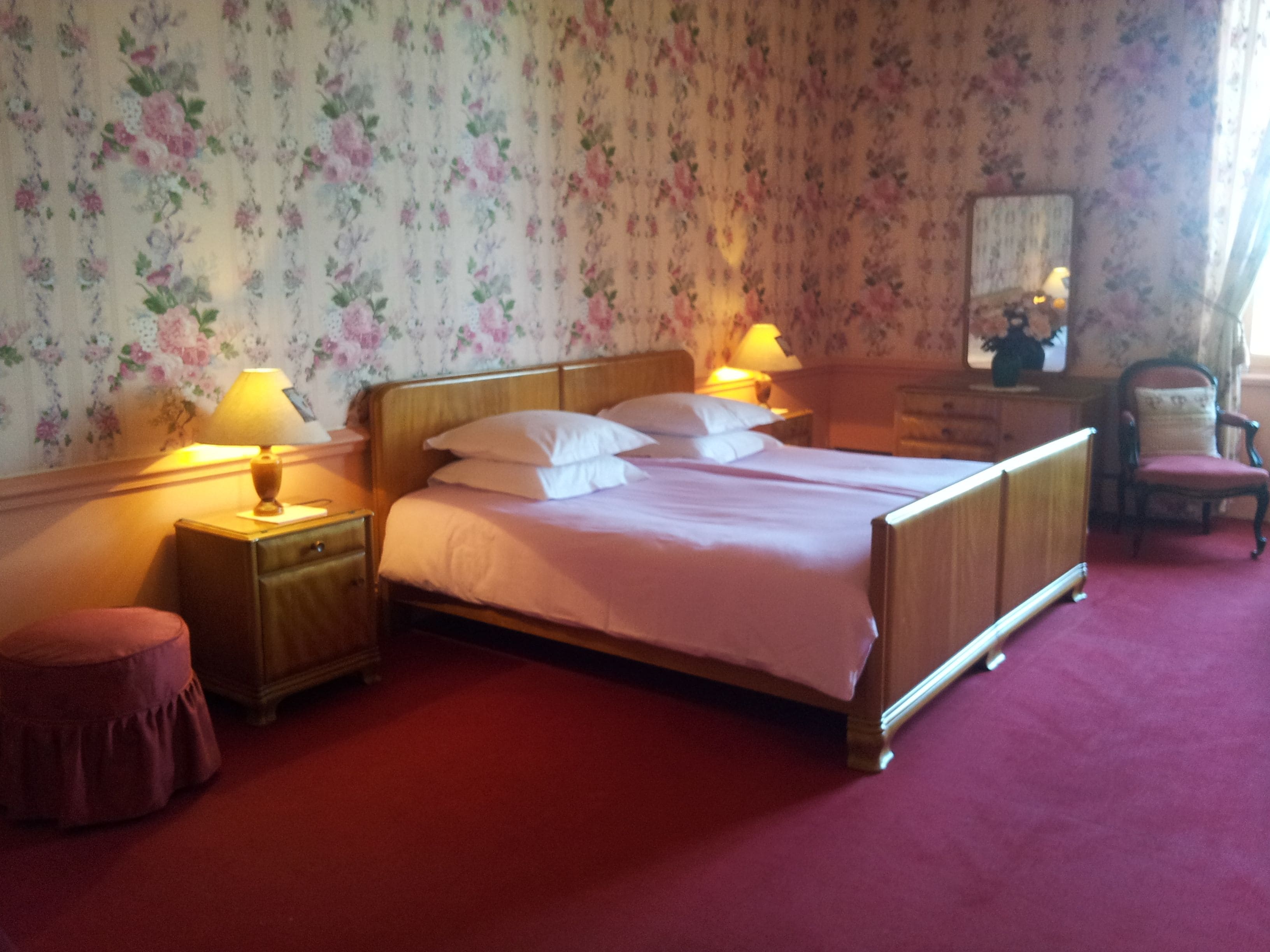 Ein Schlafzimmer mit einem hölzernen Bett, zwei Nachttischen mit Lampen, einem Sessel und blumigem Wandpapier.