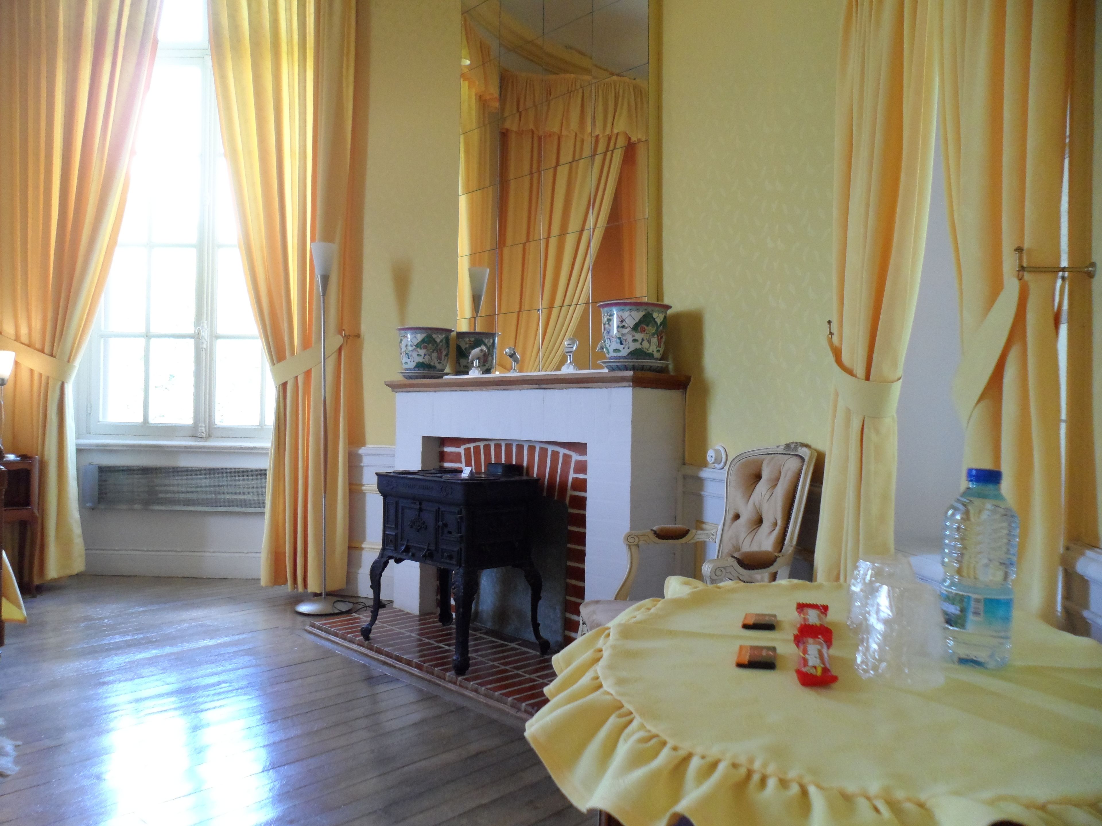 Ein Zimmer mit Kamin, einem Tisch mit gelber Tischdecke und einem Fenster mit Vorhängen.