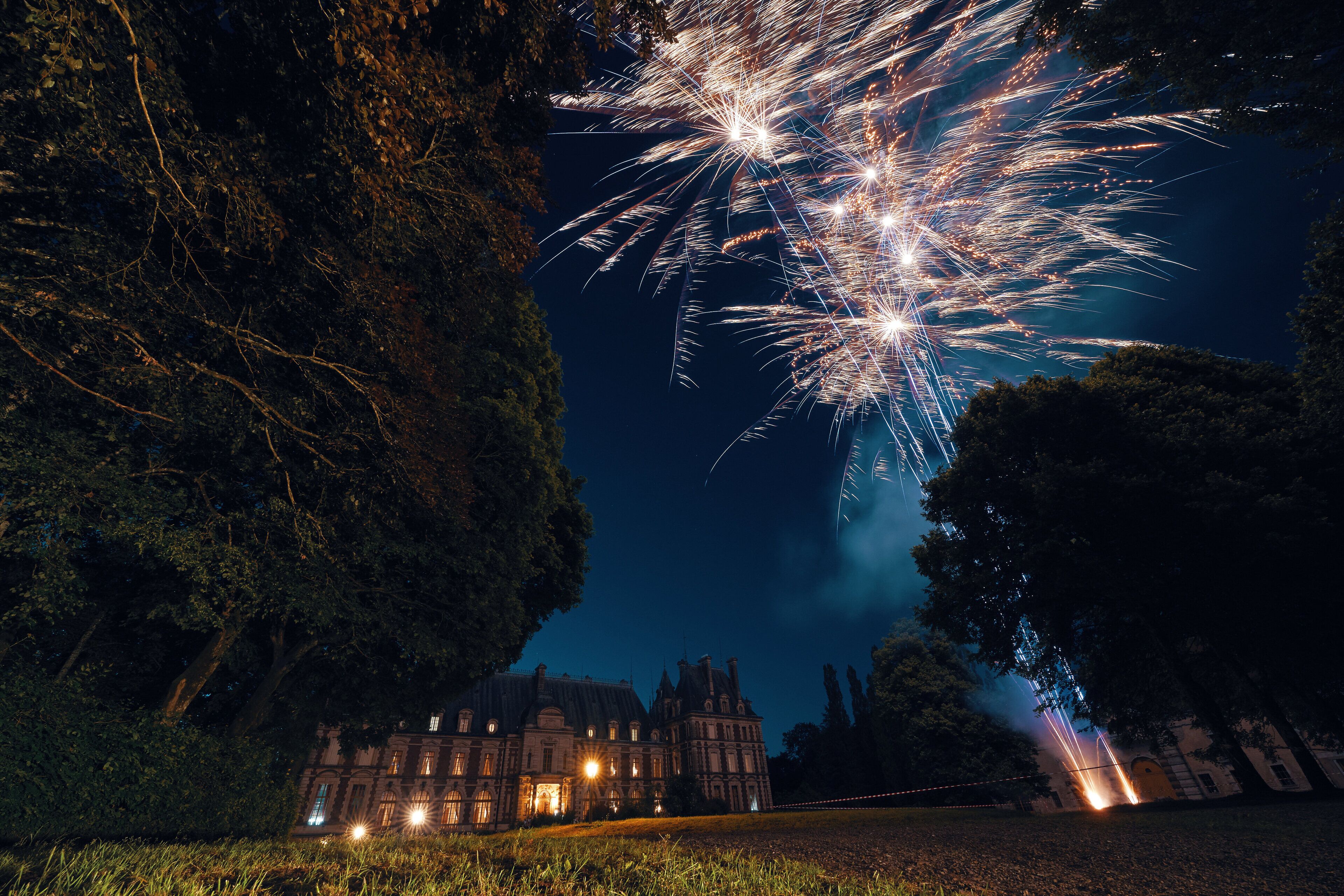 Un bâtiment historique à la façade symétrique et aux grandes fenêtres, entouré d’arbres et illuminé par des jeux de lumière de feux d’artifice.