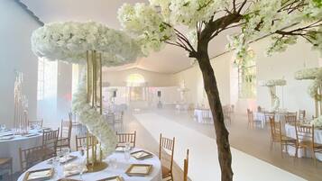Un salón de banquetes con mesas redondas dispuestas para comer, un arreglo floral blanco en el centro y un árbol artificial con flores blancas.