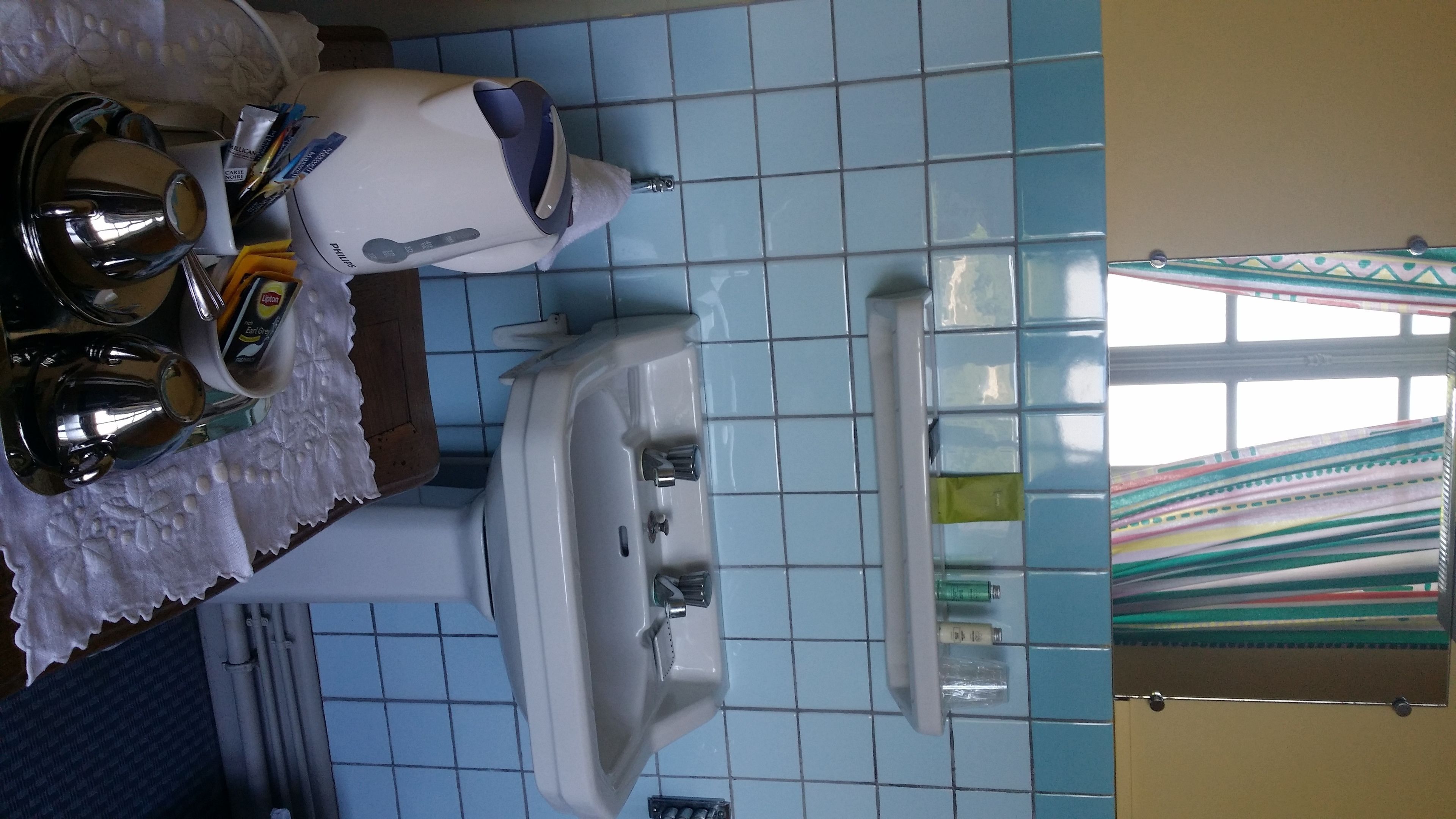 A bathroom with a white toilet, a sink, and a mirror. There is a towel rack with a white towel and a bottle of lotion. The walls are tiled in blue, and there is a window with a colorful curtain.