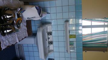 A bathroom with a white toilet, a sink, and a mirror. There is a towel rack with a white towel and a bottle of lotion. The walls are tiled in blue, and there is a window with a colorful curtain.