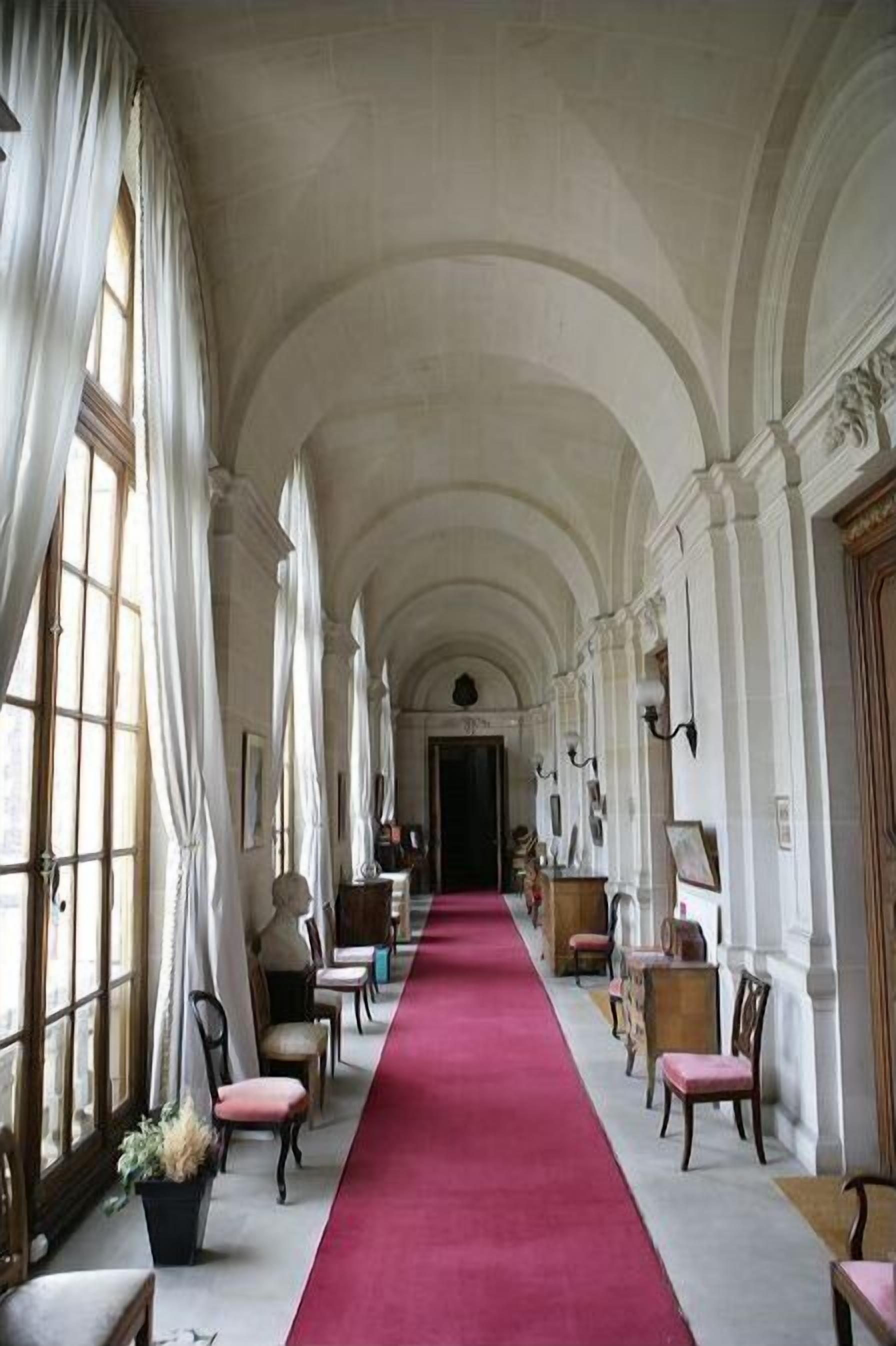 A long hallway with a red carpet, arched ceilings, and large windows with curtains.
