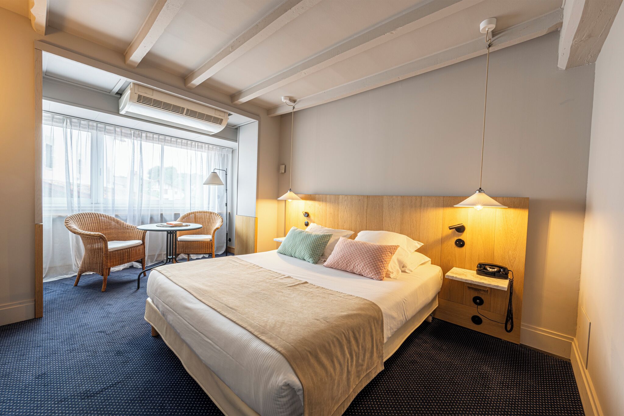 A hotel room with a large bed, two bedside tables, a chair, a small table, and a window with curtains.