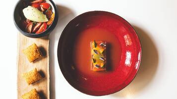 A red bowl with a clear liquid and a cube of food, and a smaller bowl with a slice of cheese and tomatoes on a wooden board.