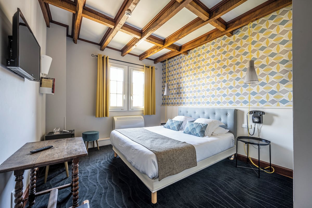 A hotel room with a bed, a desk, a chair, a telephone, and a window with curtains.