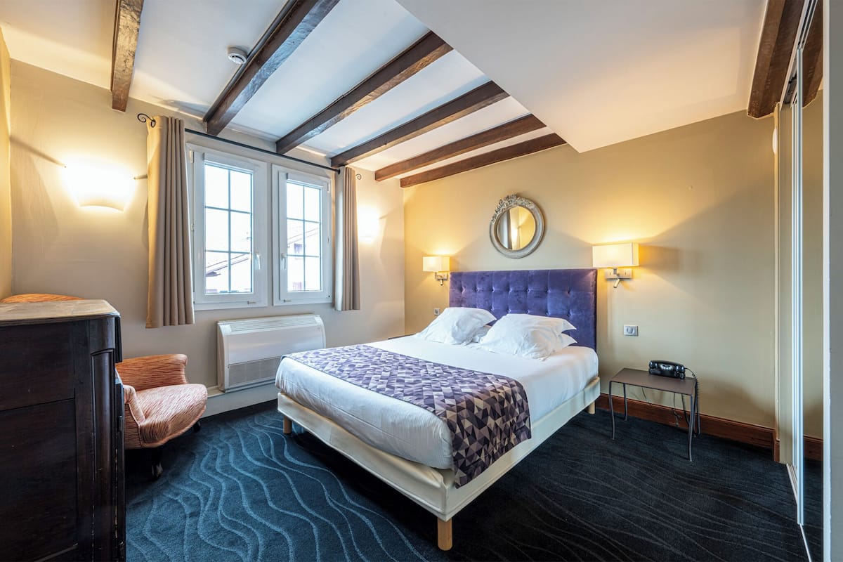 A hotel room with a large bed, a dresser, a chair, a window with curtains, a wall-mounted mirror, and two wall sconces.