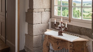 A room with a stone pillar, a large window with a view of the countryside, and a table with a candle arrangement.