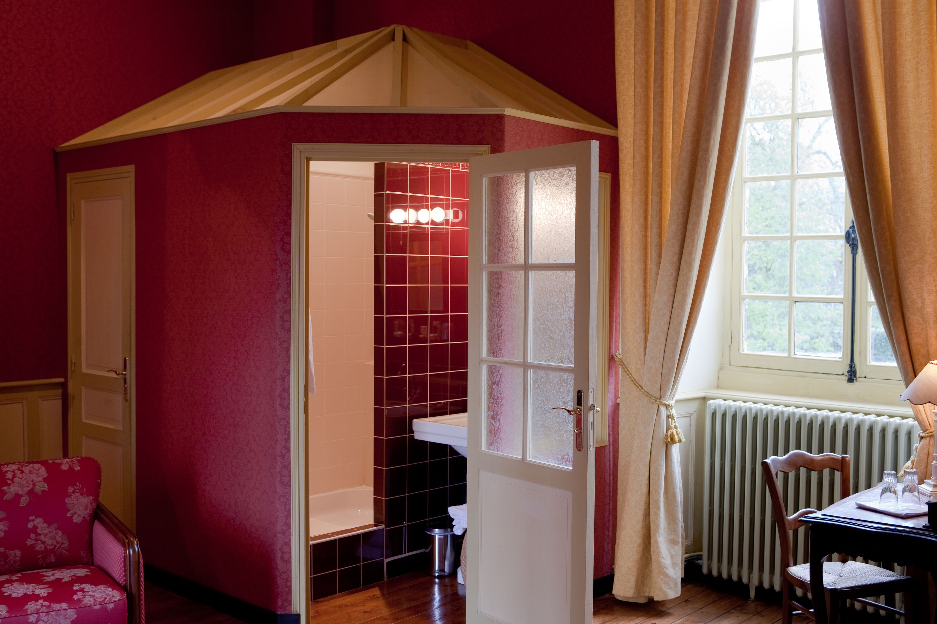 A room with a red wall, a white door leading to a bathroom, a wooden table with chairs, and a window with curtains.