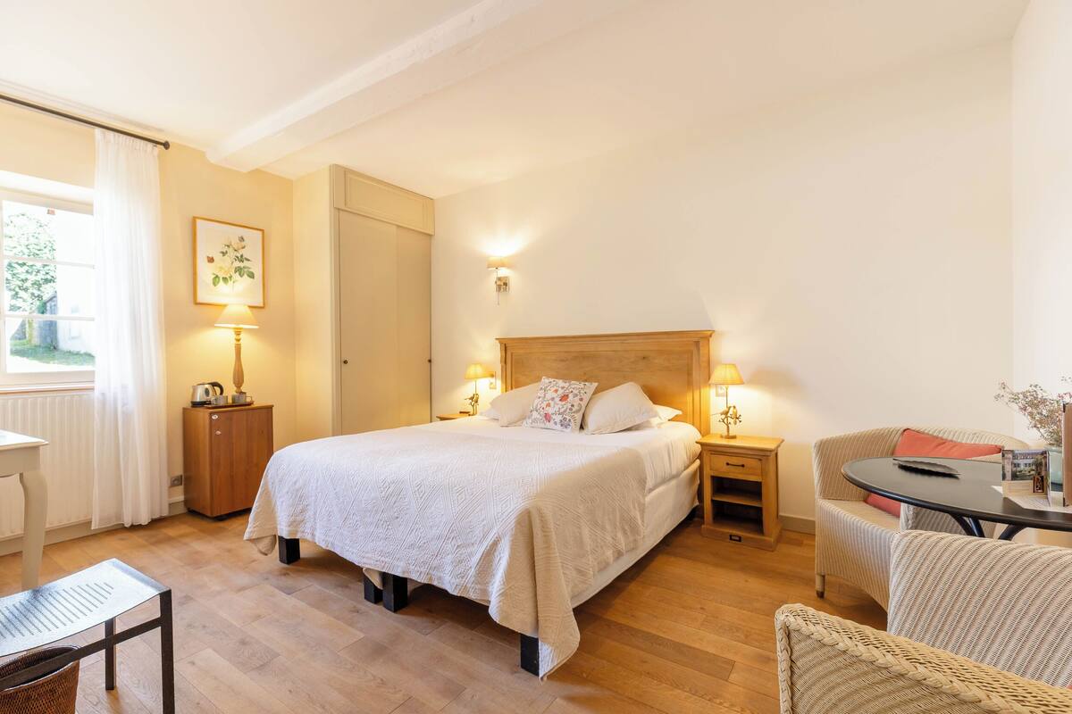 A hotel room with a bed, bedside tables, a chair, a small table, and a window with curtains.