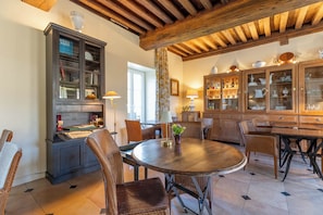 A dining area with a round table, wicker chairs, a dark cabinet with glass doors, and wooden ceiling beams. - Villa Louise (Aloxe-Corton)