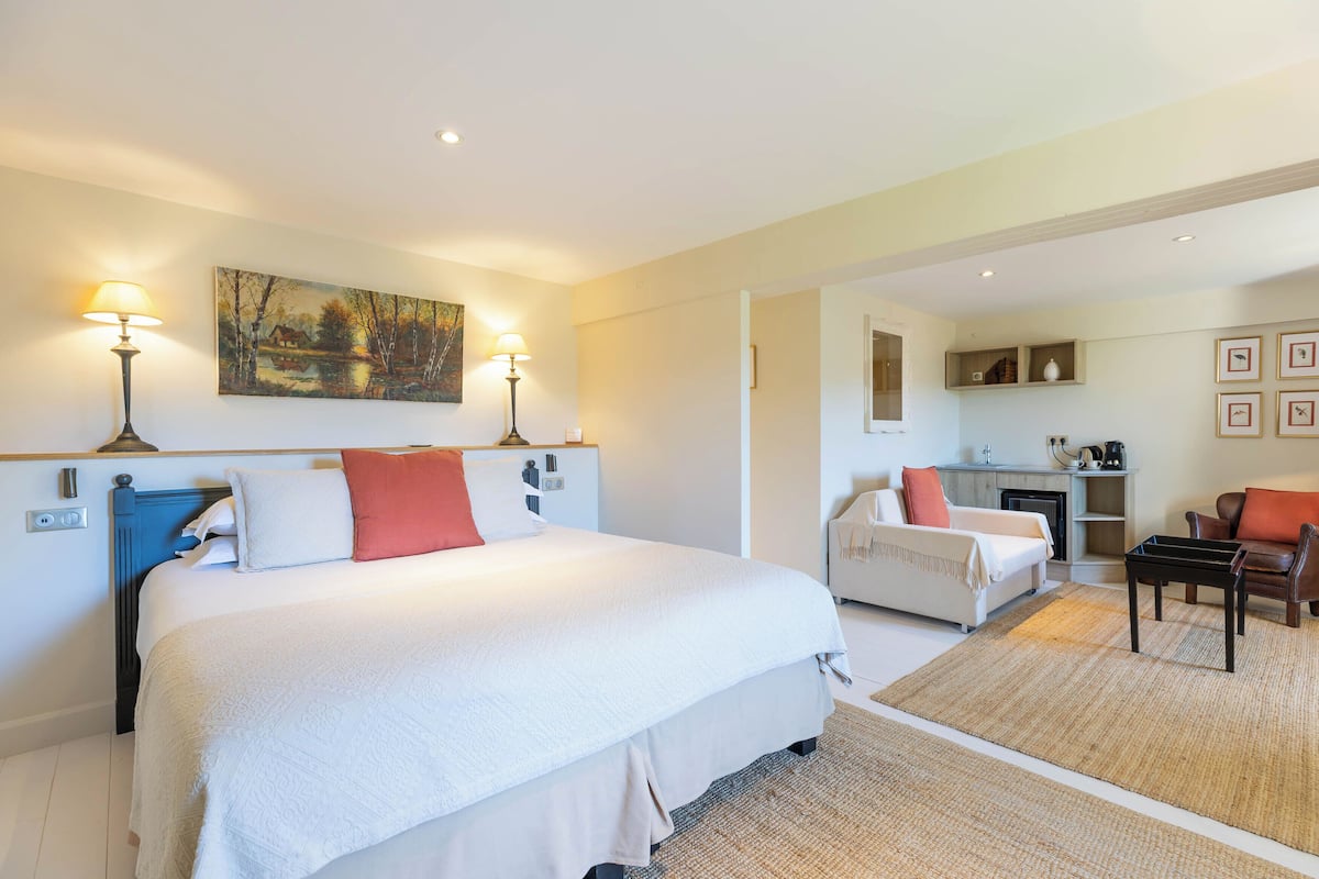 A hotel room with a large bed, two bedside tables, a desk, a chair, a sofa, a fireplace, and a painting on the wall.