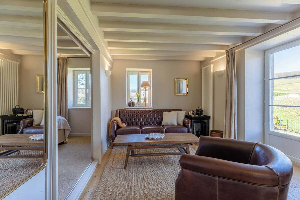 A room with a brown leather sofa, a wooden coffee table, a large window with a view, and a mirror on the wall.