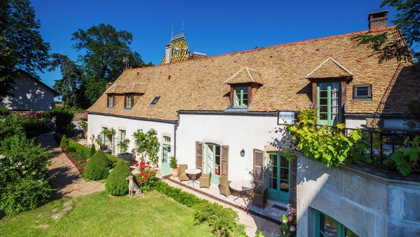 A two-story house with a brick roof, green shutters, and a small garden with a pathway leading to the entrance. - Villa Louise (Aloxe-Corton)