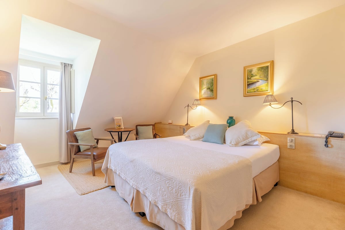 A bedroom with a large bed, two bedside lamps, a chair, a small table, and a painting on the wall.