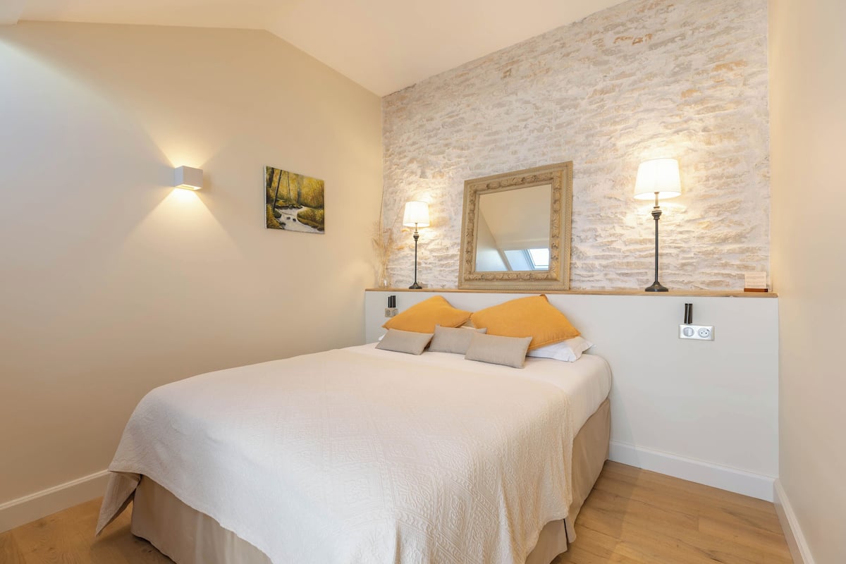 A bedroom with a bed, two wall-mounted lamps, a painting, and a stone wall.