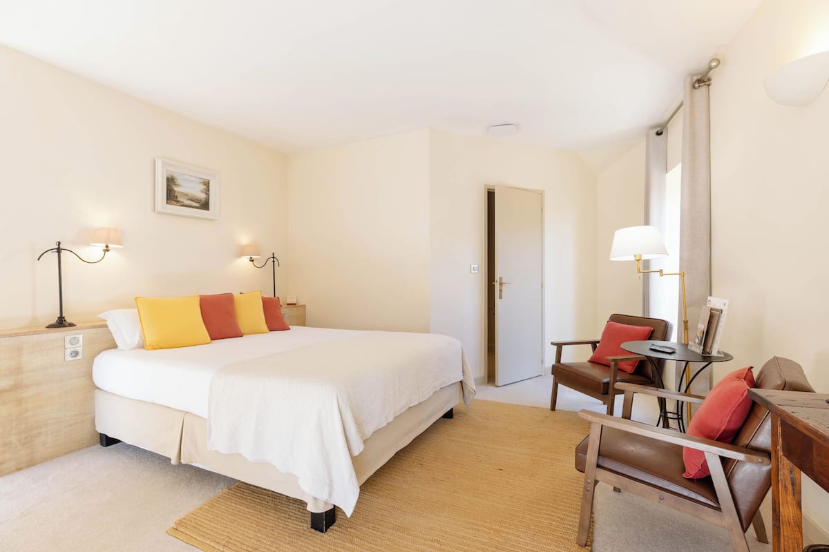 A hotel room with a bed, bedside tables, a desk, chairs, a lamp, and a door.