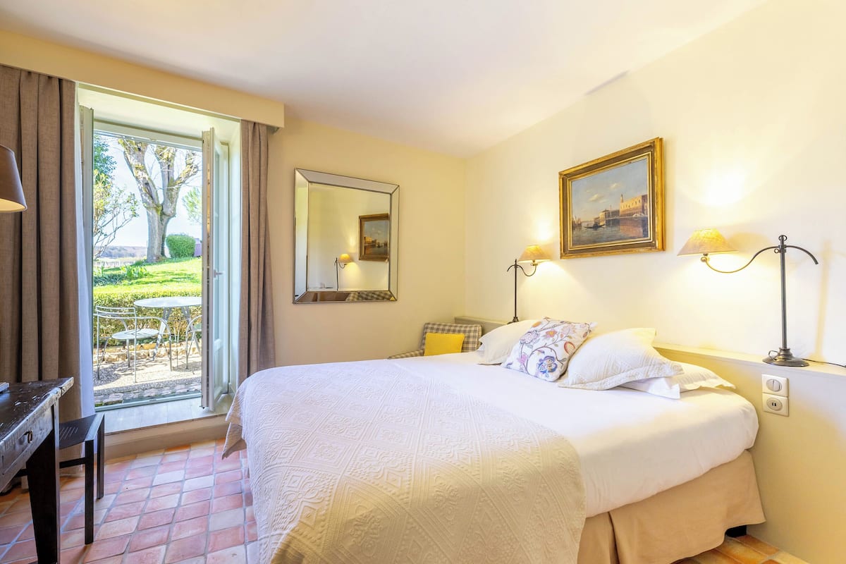 A hotel room with a bed, a desk, a chair, a window with curtains, a mirror, a painting, and a lamp.