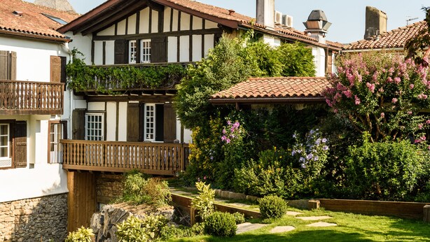 A traditional half-timbered house with a balcony, surrounded by lush greenery and flowering plants. - Hôtel Arraya (Sare)