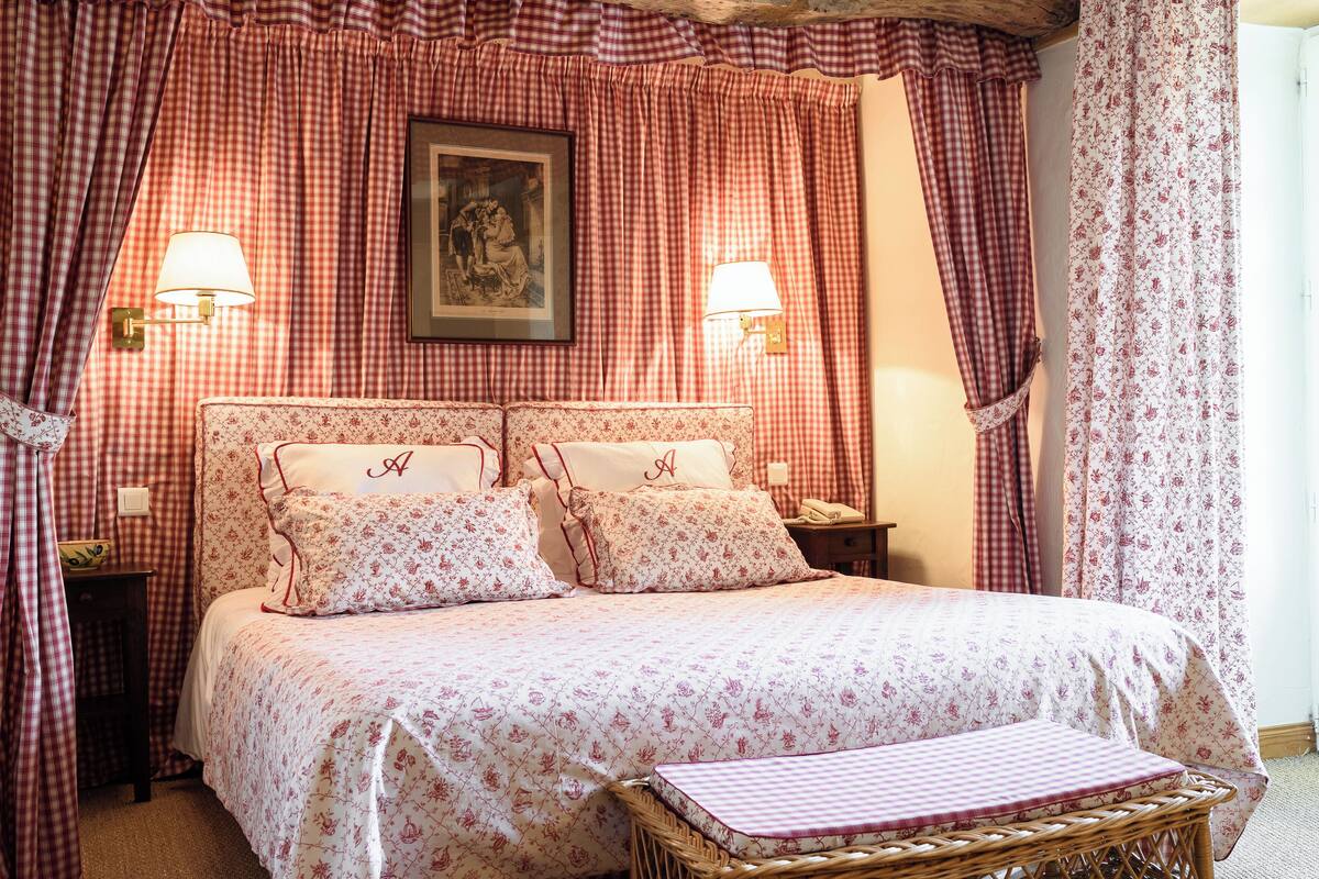 A bedroom with a checkered bedspread, two bedside lamps, a wicker bench, and a framed picture on the wall.