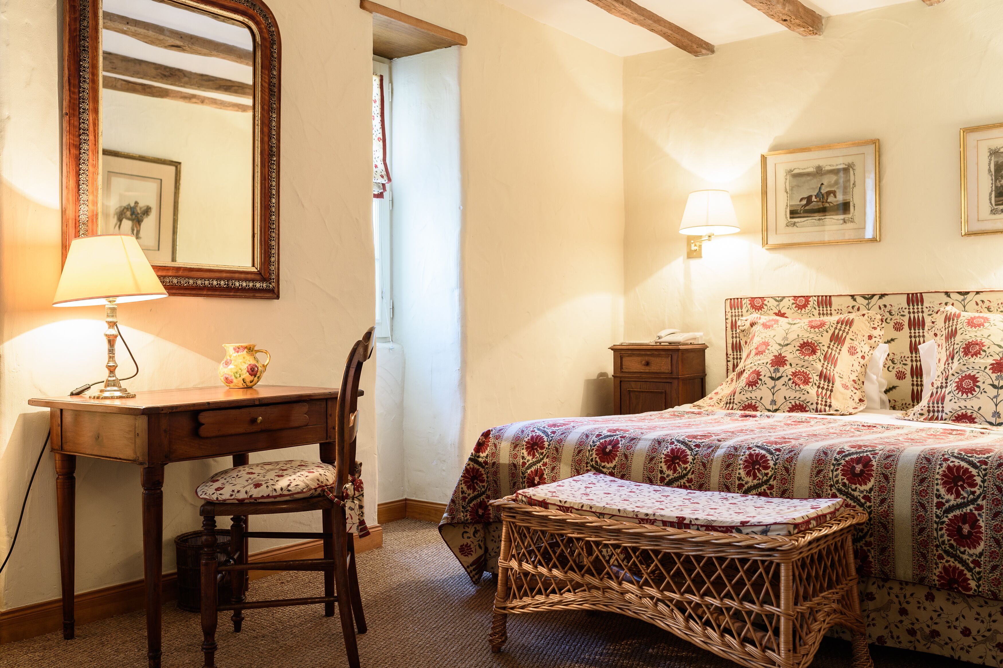 A bedroom with a bed, a desk with a lamp, a chair, a wicker bench, a mirror, and a wall with framed pictures.