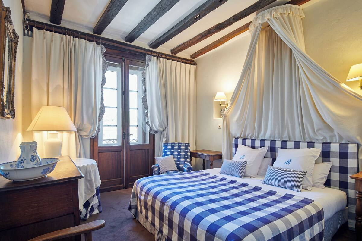 A bedroom with a checkered bedspread, a canopy bed, a wooden chair, a table with a lamp, and a window with curtains.