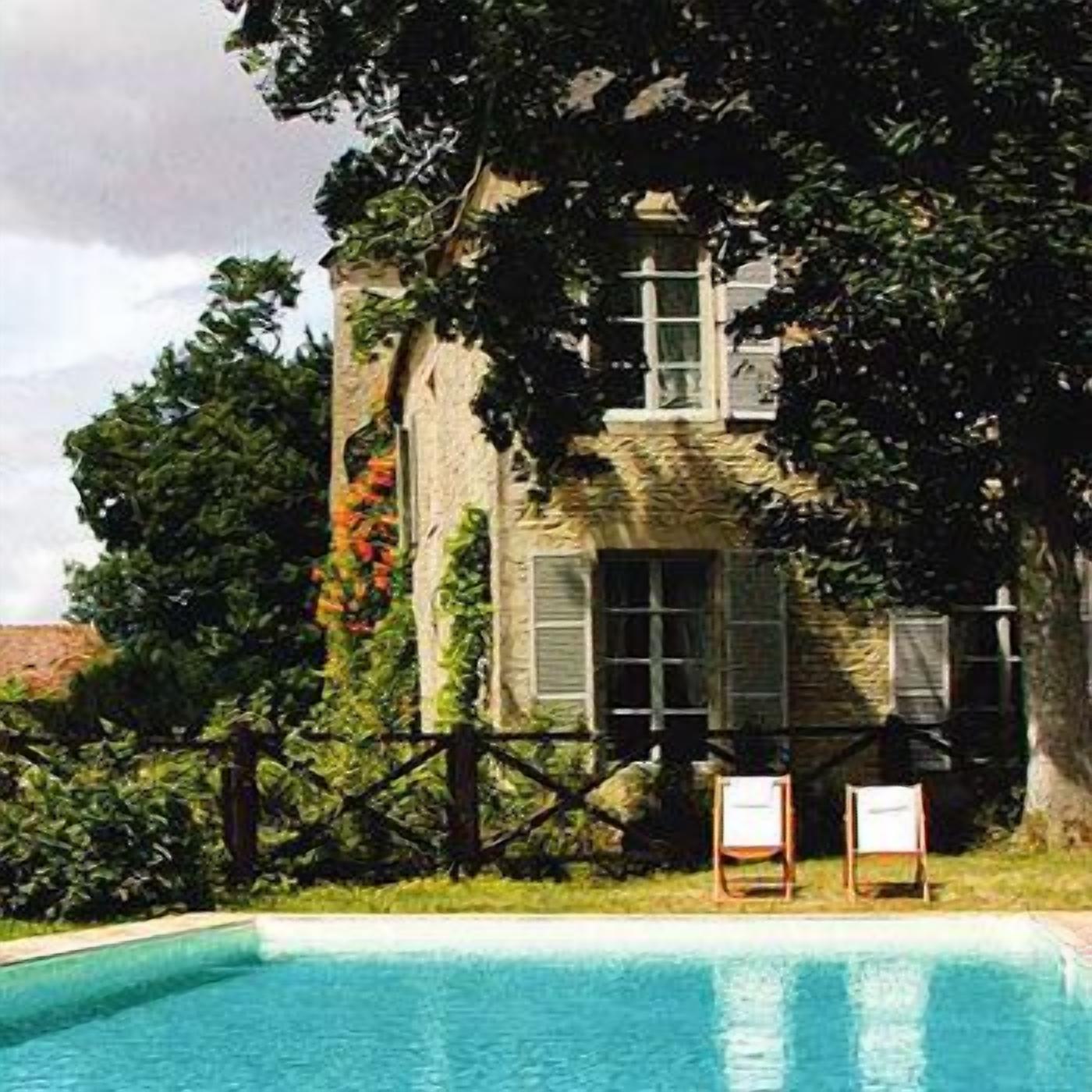 Uma piscina com duas espreguiçadeiras, um edifício de pedra com persianas e vegetação exuberante.