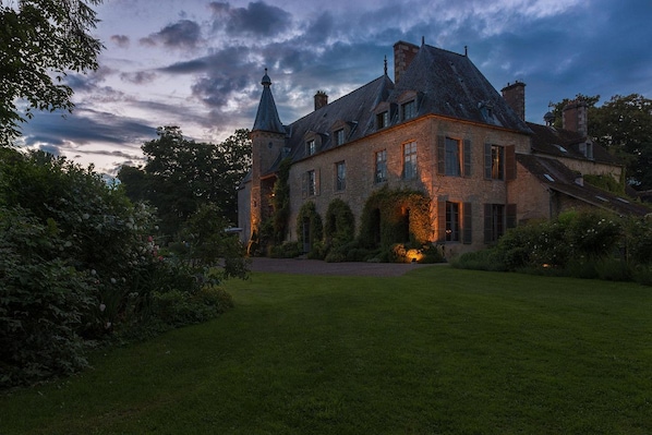 Un edificio grande e histórico con una torre destacada, rodeado de jardines cuidados y un césped impecable.