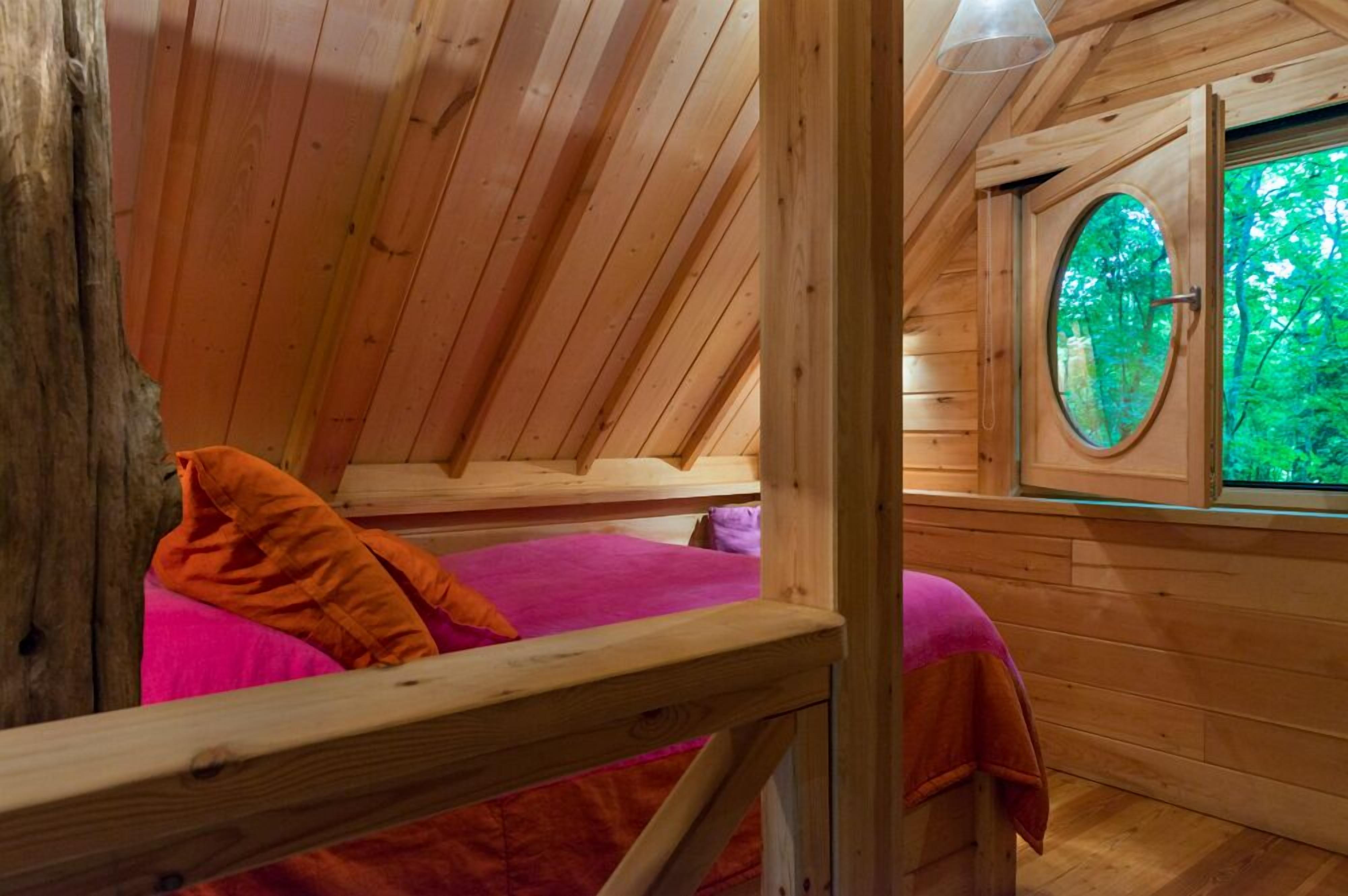 Une cabane en bois avec un lit en mezzanine, une fenêtre donnant sur la verdure et une rambarde en bois.