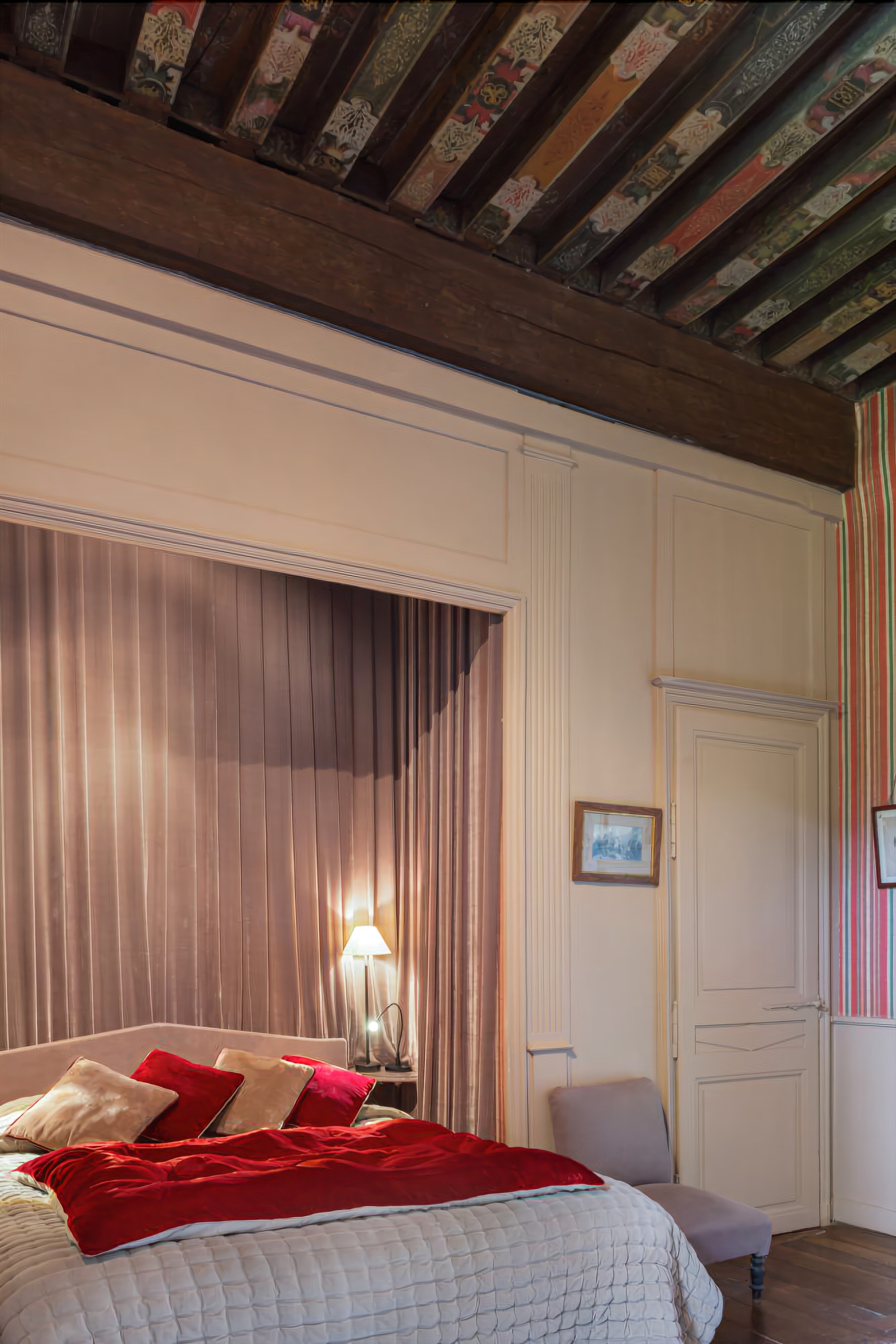 Une chambre à coucher avec un lit, des rideaux, une chaise, une lampe et une porte.