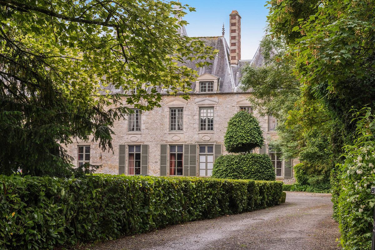 Un bâtiment historique en pierre, doté d’une façade symétrique, d’une entrée centrale et d’un chemin bordé de haies, bien entretenu, qui mène à l’entrée.
