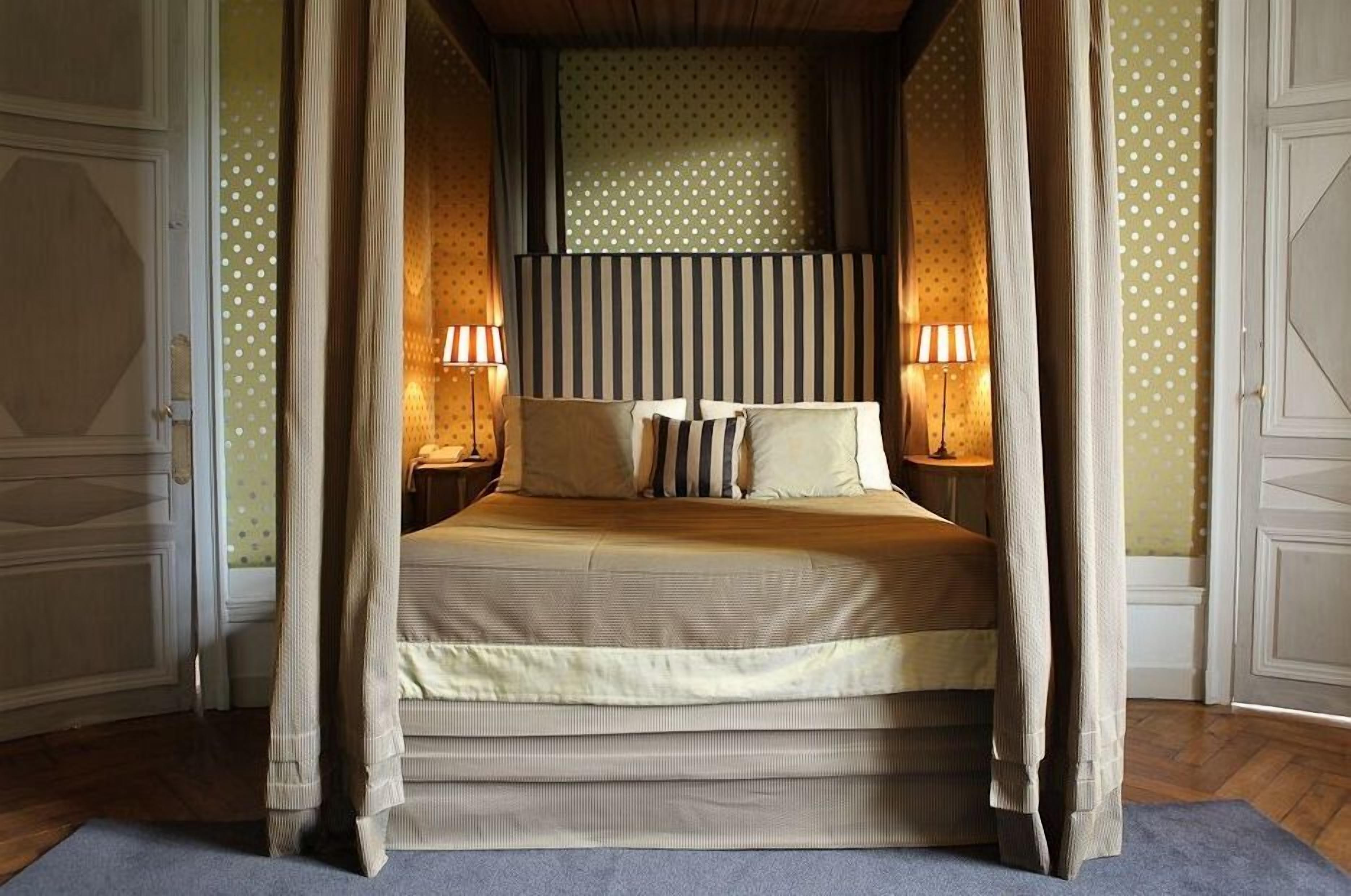 A four-poster canopy bed with striped headboard, two bedside lamps, and a patterned wallpaper.
