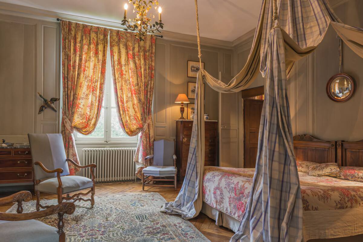 Een slaapkamer met hemelbed, een stoel, een bureau en een groot raam met gordijnen.