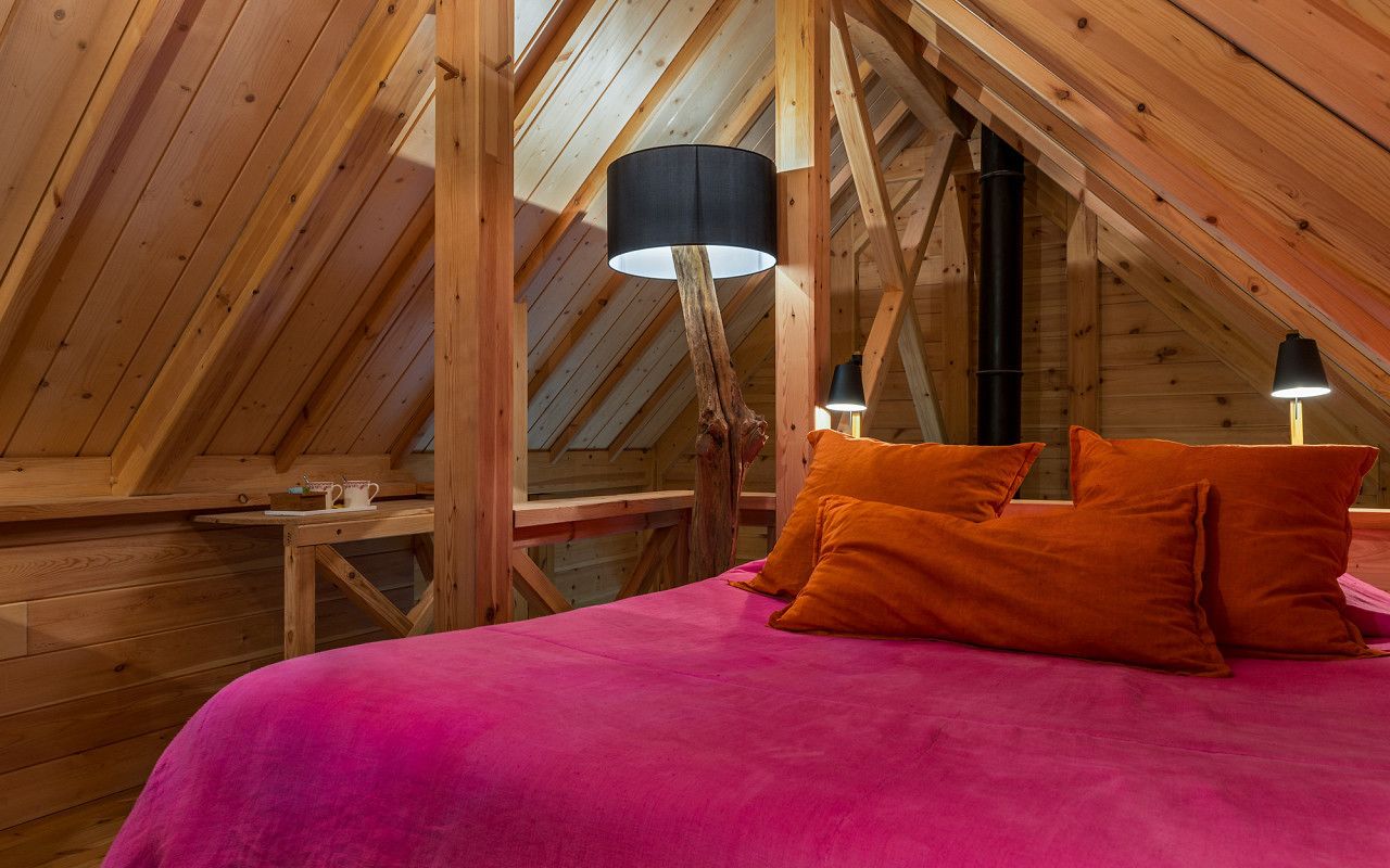 A cozy attic bedroom with a sloped ceiling, wooden beams, a bed with orange pillows, and two table lamps.
