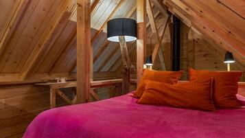 A cozy attic bedroom with a sloped ceiling, wooden beams, a bed with orange pillows, and two table lamps.