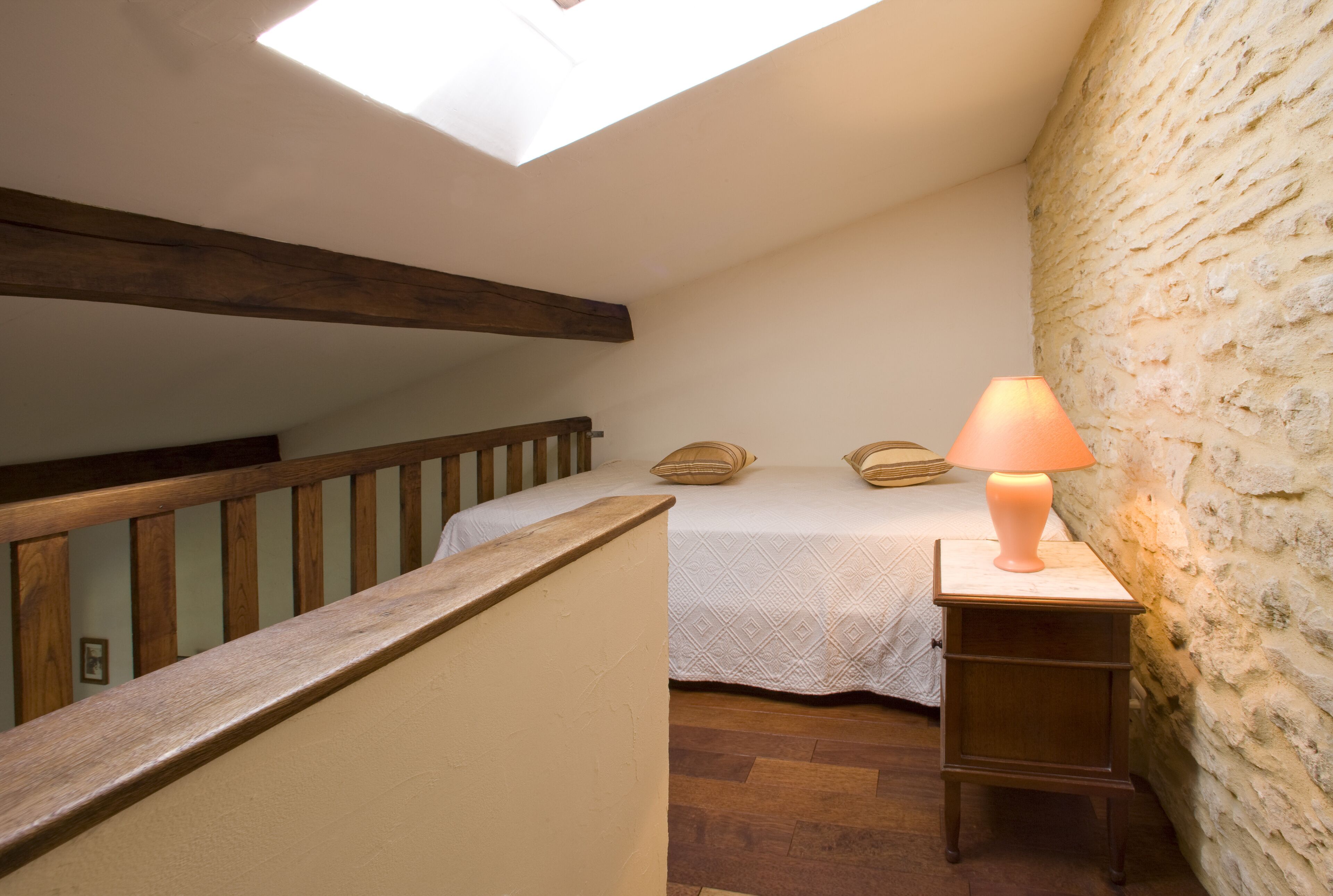 A bedroom with a skylight, a bed with pillows, a bedside table with a lamp, and a wooden staircase.