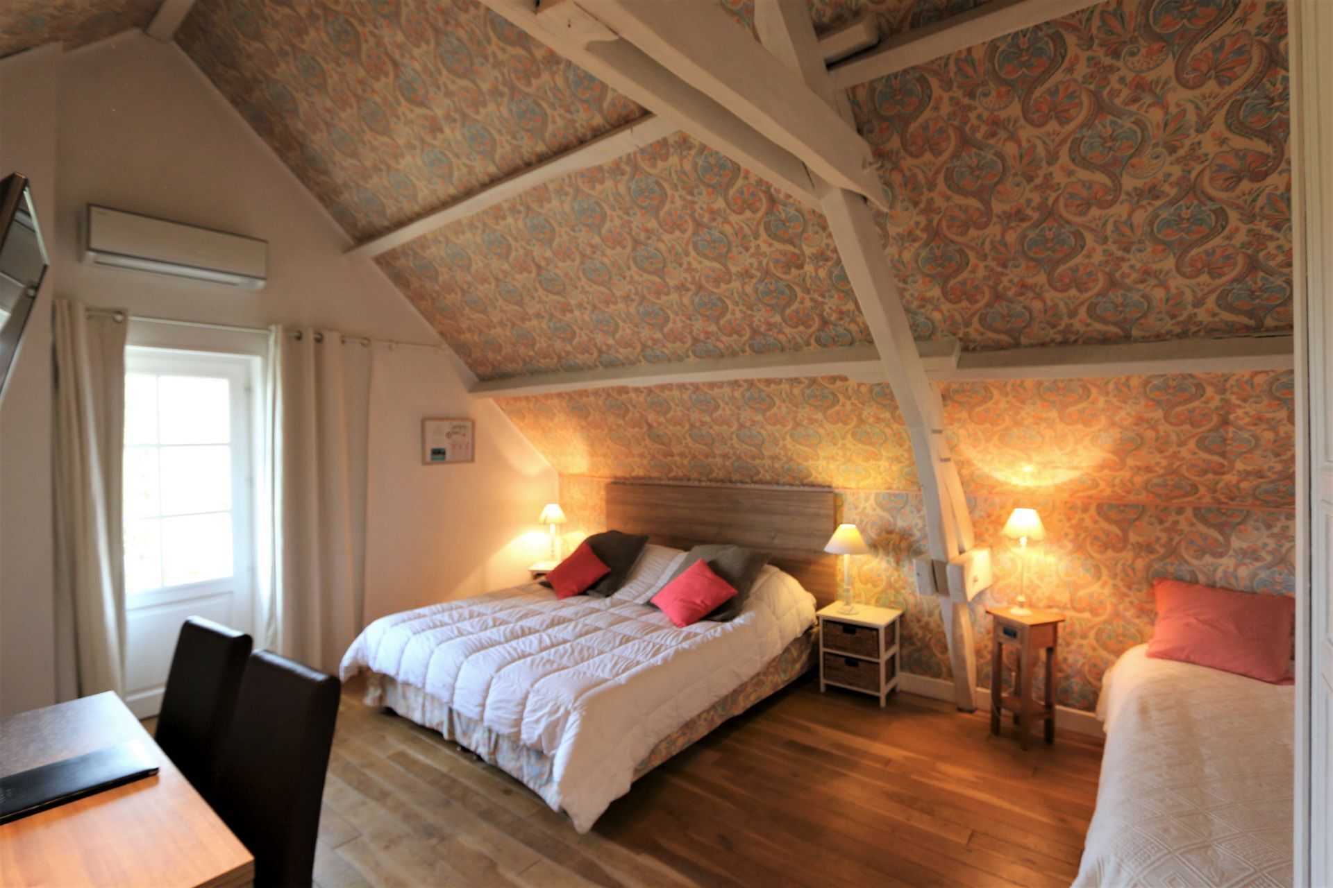 Un dormitorio con techo de madera, papel tapiz estampado, una cama con almohadas, mesitas de noche con lámparas y suelo de madera.