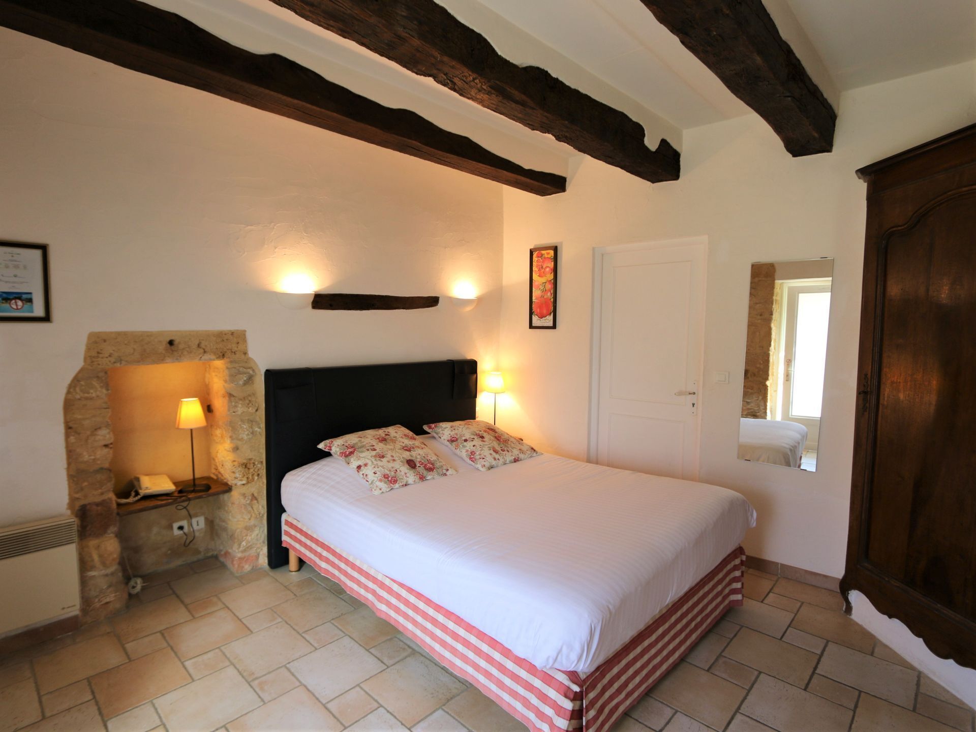 A bedroom with a bed, a stone wall, a wooden ceiling beam, a door, and a mirror.