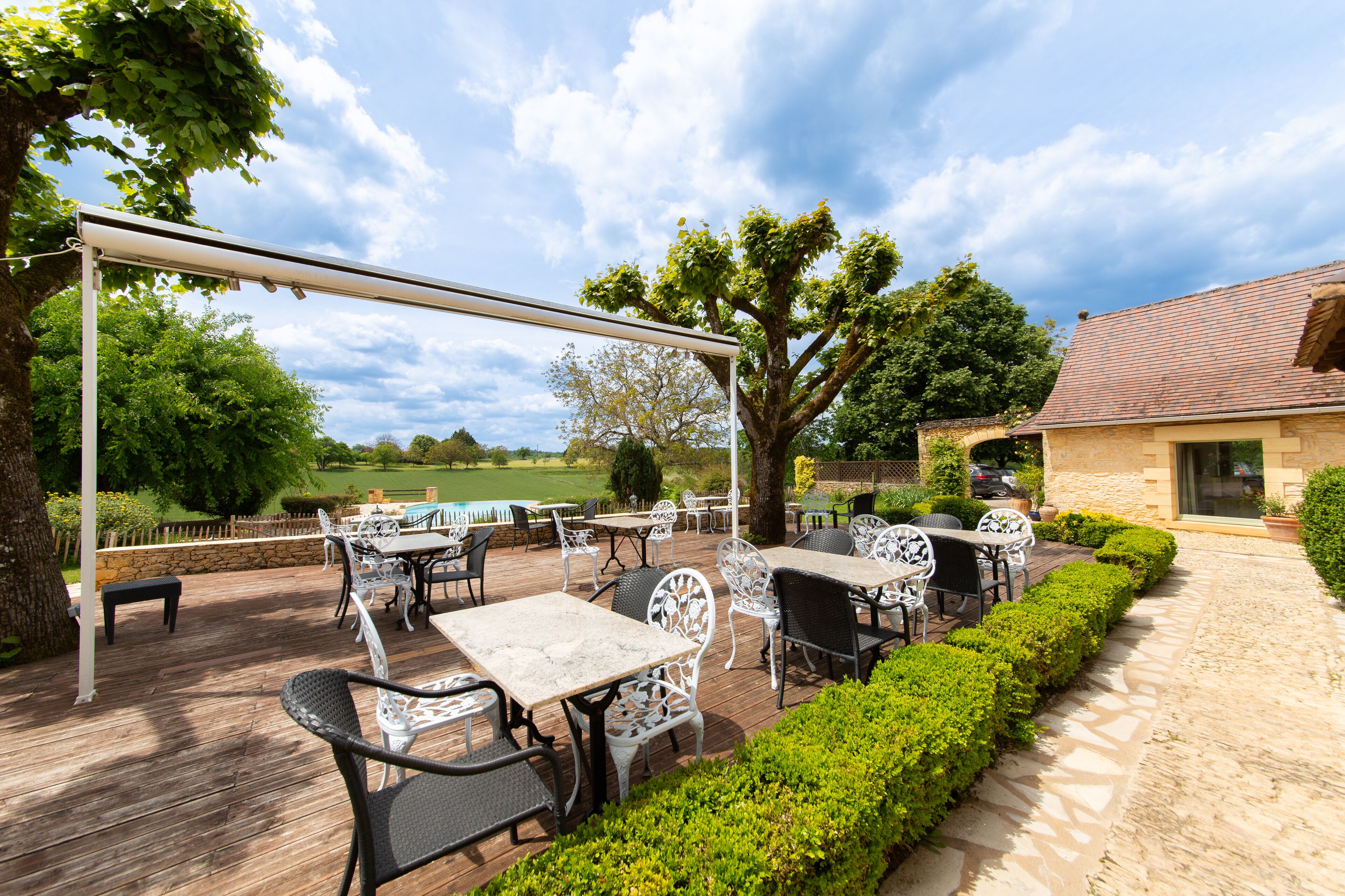 An outdoor patio with tables and chairs, surrounded by greenery and a stone building.