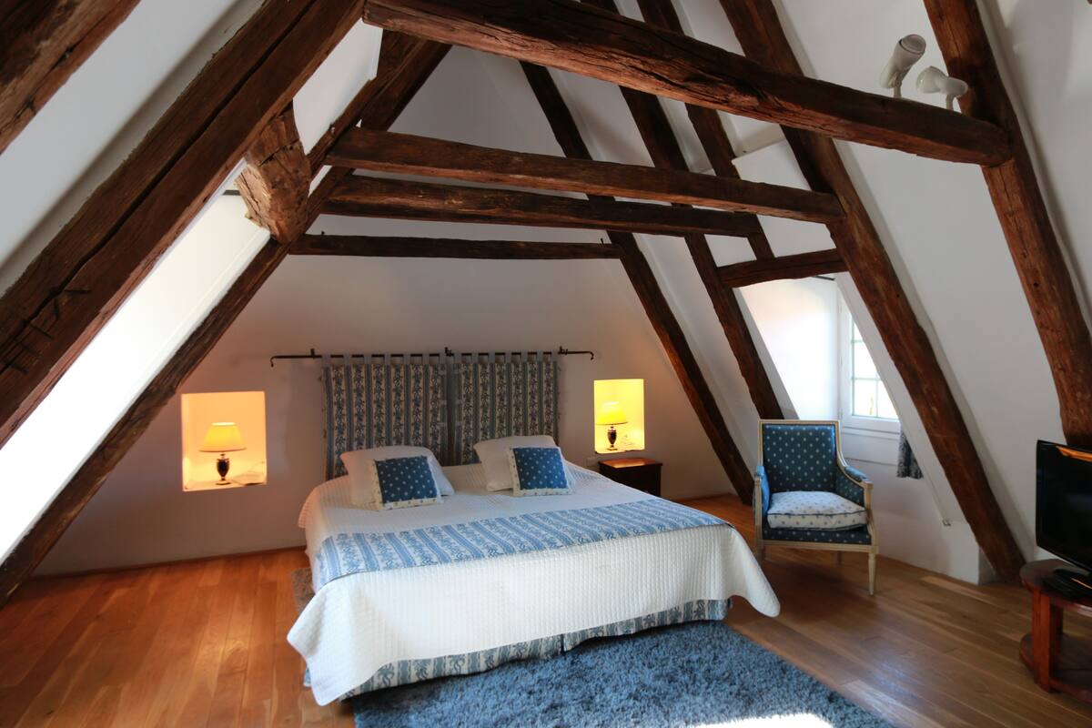 A bedroom with a sloped ceiling, wooden beams, a bed with a blue and white bedspread, two bedside lamps, a chair, and a television.