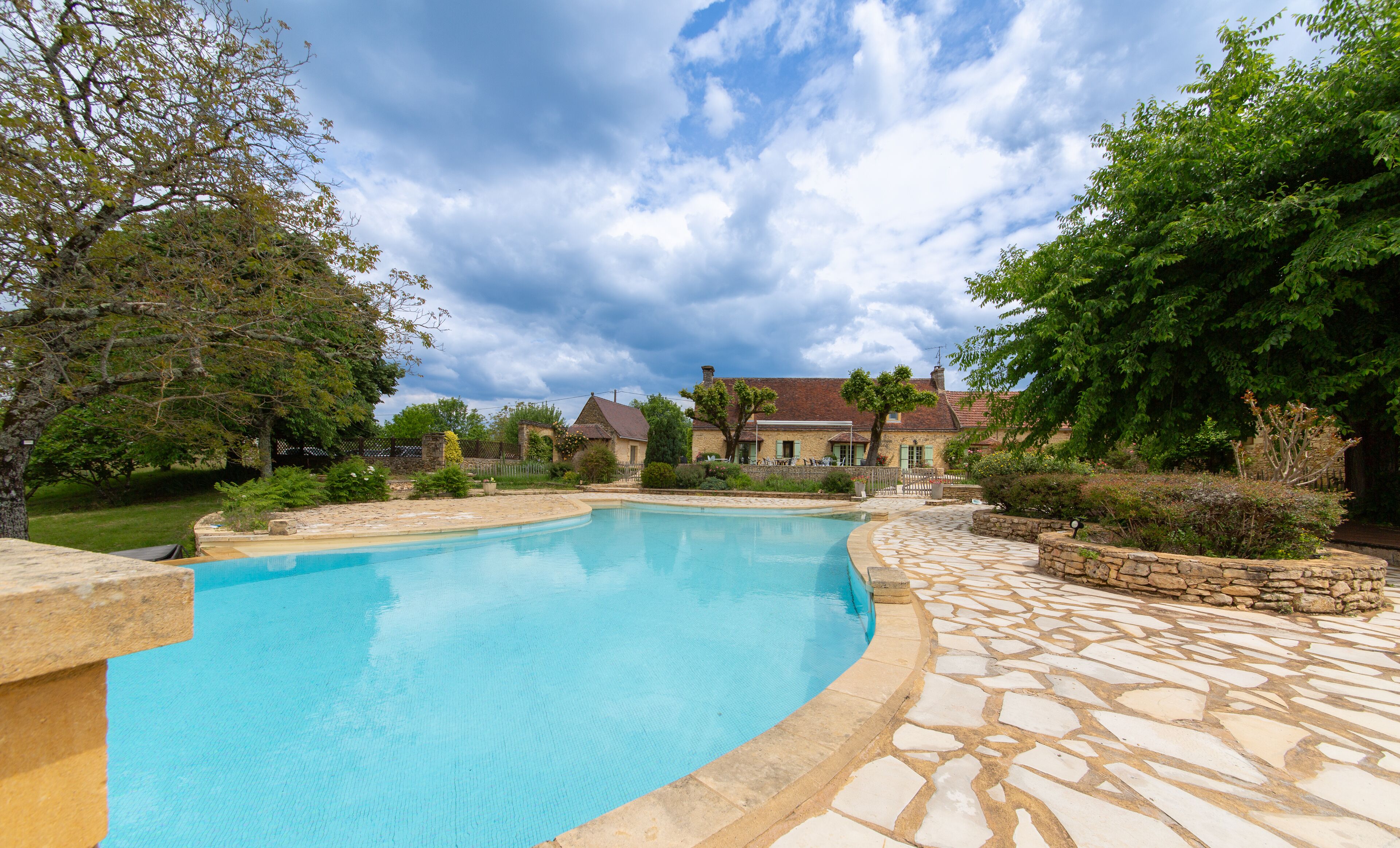 Une piscine aux bords recouverts de pierres, entourée d’un jardin et d’une maison à toit rouge.