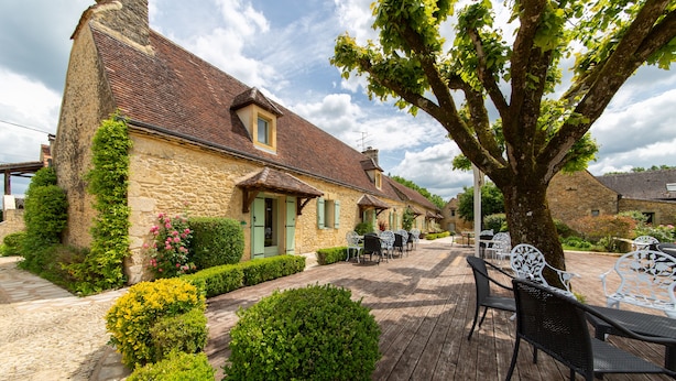 A stone building with a tiled roof, outdoor seating, and a tree in the foreground. - H ôtel de la Ferme Lamy (Meyrals)