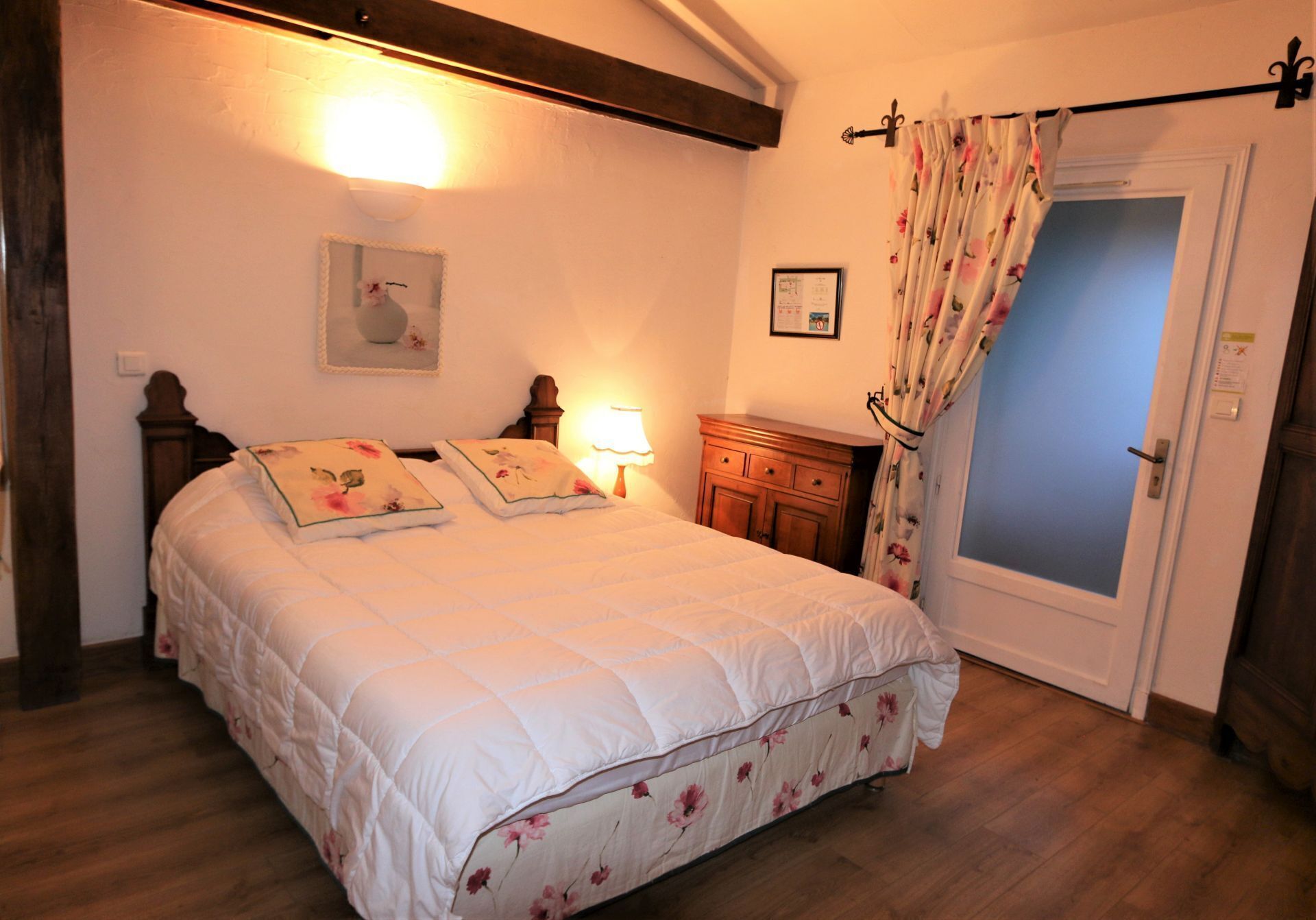 Un dormitorio con una cama grande, cabecera de madera, una mesita de noche con lámpara, cómoda y una puerta con cortina.